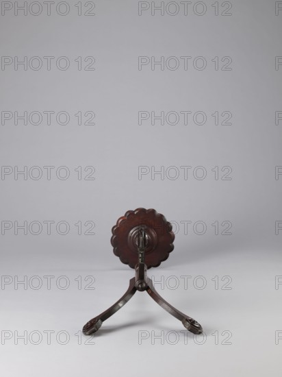 Side table, TABLE with a top in the form of a round saucer mounted on a turned stem forked into a tripod. The top has cut-out recesses for a jug and eight glasses. The central stem is in the form of a baluster with carved acanthus leaves. Legs made of channel-shaped slats, topped with volutes at the bottom, with turned knobs applied on the sides. 1820–1840, England. Furniture; table. Height 100.5 cm, width 39 cm, depth 39 cm., A wooden stand with a dark brown finish is centered on a light gray surface against a pale gray background; the stand has a round scalloped top element and two curved legs ending in carved foot pieces, all in shades of dark brown.
