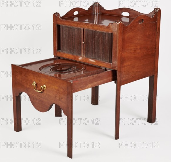 Bedside table with toilet, 4th quarter of the 18th century, England. Bedstand with toilet: tambour door in upper section and a pull-out section below with a circular opening and lid. Supported by four angular stand-up legs; front legs divided diagonally in half, the front half connected to the front wavy-cut board of the pull-out section at the bottom. Pull-out handle of gilded brass. Opening cover turned, recessed in the center, with a round handle. Front and side walls of the body have wavy-cut tops with oval openings as handles. A blind of two parts slides sideways, with small brass knobs. Function: piece of furniture for hygiene. Dimensions: height 82 cm, width 59.2 cm, depth 45 cm., A wooden piece of furniture with a warm brown finish standing on four straight legs, featuring a lower pull-out section with a scalloped apron and a brass handle, and an upper compartment with a pair of vertically ribbed panel doors set into a framed box with decorative cutouts along the top edges; the...