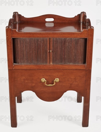Bedside table with toilet, 4th quarter of the 18th century, England. Bedstand with toilet: upper section with a two-part sliding tambour door with small brass knobs; below is a pull-out section with a circular opening for a bedside chamber pot with a lid. Supported by four angular stand-up legs; front legs divided diagonally in half, the front half connected to the front, wavy-cut board of the pull-out section at the bottom. Pull-out handle of gilded brass. Opening cover turned, recessed in the center with a round handle. Front and side walls of the body have wavy-cut tops with oval openings handles. Function: piece of furniture for hygiene. Dimensions: height 82 cm, width 59.2 cm, depth 45 cm., A wooden piece of furniture with a reddish-brown finish standing on four straight legs, featuring a raised tray-like top with curved cutouts and a small oval handle opening, a central compartment with a pair of vertically ribbed sliding doors in a darker brown, and a lower drawer with a brass-c...