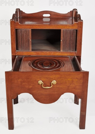 Bedside table with toilet, 4th quarter of the 18th century, England. Bedstand with toilet, with a tambour door in the upper section and a pull-out section below with a circular opening and a lid. Supported by four angular stand-up legs; the front legs divided diagonally in half, the front half connected to the front, wavy-cut board of the pull-out section at the bottom. Pull-out handle of gilded brass. Opening cover turned, recessed in the center, with a round handle. Front and side walls of the body have wavy-cut tops with oval openings handles. A blind consisting of two parts that slide sideways, with small brass knobs. A piece of furniture for hygiene; materials and style consistent with 18th-century English furniture. Height 82 cm, width 59.2 cm, depth 45 cm., The image shows a wooden piece of furniture finished in warm brown tones with a glossy surface, featuring a top tray-like section with a cutout handle and raised edges, an open shelf area with sliding ribbed panels on each si...