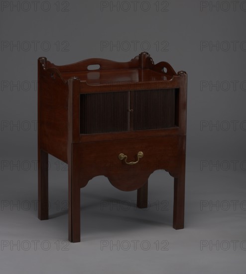 Bedside table with toilet, with a tambour door in the upper section and a pull-out section below with a circular opening for a chamber pot with a lid. Supported by four angular stand-up legs; front legs divided diagonally in half, the front half connected to the front, wavy-cut board of the pull-out section at the bottom. Pull-out handle of gilded brass. Opening cover turned, recessed in the center, with a round handle. Front and side walls of the body have wavy-cut tops with oval openings as handles. A blind consisting of two parts that slide sideways, with small brass knobs. 4th quarter of the 18th century, England, a piece of furniture for hygiene; height 82 cm, width 59.2 cm, depth 45 cm., A wooden piece of furniture with a dark brown finish standing on four straight legs, featuring a rectangular top with curved raised sides and cutout handles, a recessed middle section with sliding tambour-style doors, an open lower drawer front with a brass-colored handle, and a scalloped apron; ...