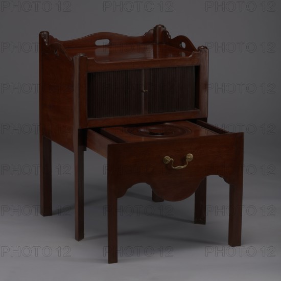 Bedside table with toilet, hygiene, BEDSTAND WITH TOILET, with a tambour door in the upper section and a pull-out section below with a circular opening for a bedside chamber pot with a lid. Supported by four angular stand-up legs; the front legs divided diagonally in half, the front half connected to the front, wavy-cut board of the pull-out section at the bottom. The pull-out handle is gilded brass. The opening cover is turned, recessed in the center, with a round handle. The front and side walls of the body have wavy-cut tops with oval openings as handles. A blind consisting of two parts that slide sideways, with small brass knobs. 4th quarter of the 18th century, England, furniture for hygiene, height 82 cm, width 59.2 cm, depth 45 cm., A wooden piece of furniture with a rich brown finish standing on a group of slender legs, featuring a raised back with curved decorative cutouts and rounded corner posts, a recessed top surface, a pair of vertical slatted panels in the upper section,...