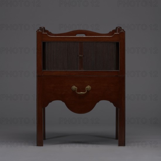 Bedside table with toilet, 4th quarter of the 18th century, England. Bedstand with toilet: tambour door in the upper section and a pull-out section below with a circular opening for a bedside chamber pot with a lid. Supported by four angular stand-up legs; front legs divided diagonally in half, the front half connected to the front, wavy-cut board of the pull-out section at the bottom. Pull-out handle of gilded brass. Opening cover turned, recessed in the center, with a round handle. Front and side walls of the body have wavy-cut tops with oval openings handles. A blind consisting of two parts that slide sideways, with small brass knobs. Function: furniture for hygiene. Dimensions: height 82 cm, width 59.2 cm, depth 45 cm., A wooden piece of furniture with a dark brown finish standing against a gray background, showing a rectangular upper compartment with a pair of vertically ridged sliding panels in a darker brown, a lower panel with a curved apron and a brass-colored handle, straight...