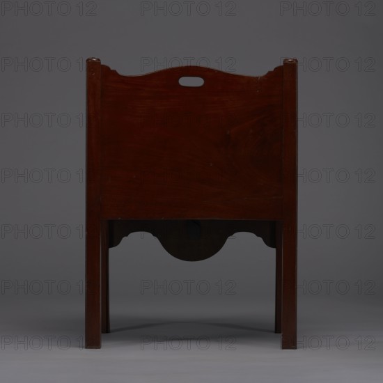 Bedside table with toilet, tambour door in the upper section and a pull-out section below with a circular opening for a bedside table with a lid. Supported by four angular stand-up legs; the front legs divided diagonally in half, the front half connected to the front, wavy-cut board of the pull-out section at the bottom. Pull-out handle of gilded brass. Opening cover turned, recessed in the center, with a round handle. Front and side walls of the body have wavy-cut tops with oval openings—handles. A blind consisting of two parts that slide sideways, with small brass knobs. 4th quarter of the 18th century, England. A piece of furniture for hygiene. Height 82 cm, width 59.2 cm, depth 45 cm., A wooden piece of furniture viewed from one side, showing a dark reddish-brown rectangular panel with a curved top edge and a centered oval cutout, supported by straight vertical legs of similar wood. The lower section has a scalloped apron with a curved central recess that creates shadow beneath it....