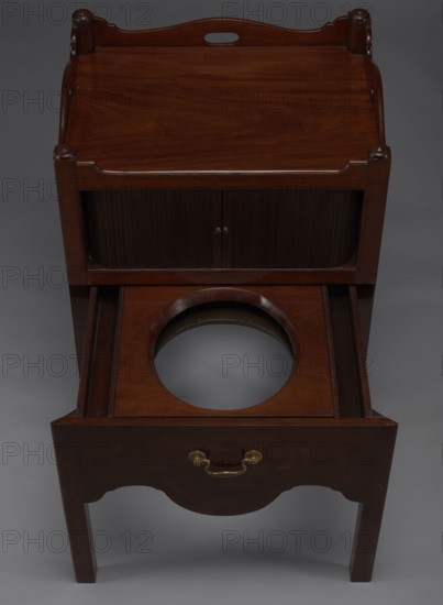 Bedside table with toilet, 4th quarter of the 18th century, England. Bedstand with toilet: tambour door in the upper section and a pull-out section below with a circular opening for a bedside chamber pot with a lid. Supported by four angular stand-up legs; front legs divided diagonally in half, the front half connected to the front, wavy-cut board of the pull-out section at the bottom. Pull-out handle of gilded brass. Opening cover turned, recessed in the center, with a round handle. Front and side walls of the body have wavy-cut tops with oval openings handles. A blind consisting of two parts that slide sideways, with small brass knobs. Function: piece of furniture for hygiene. Height 82 cm, width 59.2 cm, depth 45 cm., A wooden piece of furniture with a polished dark brown finish is shown from above, featuring a top surface with raised edges and rounded corner posts, a middle section with a pair of vertically ribbed doors, and a lower pull-out drawer opened to reveal a circular cutou...