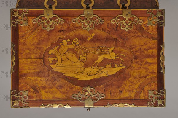 Casket, repossession provenance; sleigh, deer, hunters, hounds, riders; Baroque style; cuboid case inlaid with figural scenes, with openwork brass fittings. Front of body: scene of a deer hunt with dogs; lid: oval field showing a sleigh drawn by a deer. Inside the body, beneath the chamber floor, are three drawers middle one smaller accessible after folding down the front wall. Lid contains a compartment closed with a plate on brass hinges. Fittings cut from thick brass sheet placed on lid edges and body corners. Bottom and back wall fastened with strap hinges with decorative openwork finials visible from back of lid and at bottom of front wall. Mortice-type lock with two catches mounted on bottom of lid. Date: 1720–1740. Place of origin: Germany. Furniture — storage furniture. Dimensions: height 21.6 cm, width 42.5 cm, depth 27.4 cm., A rectangular wooden panel with a warm brown and golden-brown grain, bordered by darker brown framing and decorative brass fittings at the corners and a...