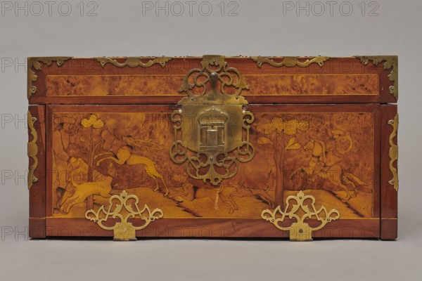 Casket, Repossession provenance. Sleigh, deer, hunters, hounds, riders. Baroque style. Cuboid case inlaid with figural scenes, with openwork brass fittings. Front wall of the body shows a deer hunt with dogs; on the lid, in an oval field, a sleigh drawn by a deer. Inside the body, under the chamber floor, are three drawers middle one smaller accessible after folding down the front wall. Lid contains a compartment closed with a plate on brass hinges. Fittings cut from thick brass sheet placed on the edges of the lid and the corners of the body. Bottom and back wall fastened with strap hinges with decorative openwork finials visible from the back of the lid and at the bottom of the front wall. Mortice-type lock with two catches mounted on the bottom of the lid. Dated 1720–1740, Germany. Furniture — storage furniture. Dimensions: height 21.6 cm; width 42.5 cm; depth 27.4 cm., A rectangular wooden chest with a warm brown and amber marbled surface, decorated with ornate brass fittings and e...