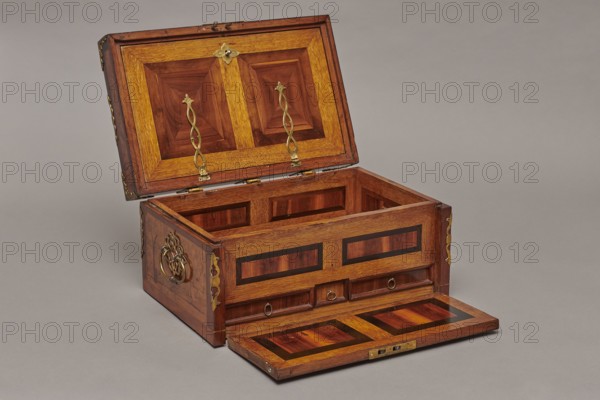 Casket by, c. 1720–1740, Germany. Baroque cuboid case inlaid with figural scenes and openwork brass fittings. Front shows a deer hunt with dogs; lid has an oval field depicting a sleigh drawn by a deer. Interior: beneath the chamber floor are three drawers middle smaller accessible after folding down the front wall; lid contains a compartment closed with a plate on brass hinges. Thick brass fittings on lid edges and body corners; bottom and back wall fastened with strap hinges with decorative openwork finials visible from back of lid and bottom of front wall. Mortice-type lock with two catches mounted on bottom of lid. Furniture — storage casket. Dimensions: H 21.6 cm, W 42.5 cm, D 27.4 cm., An open wooden chest with a hinged lid and a drop-down front panel sits on a neutral gray surface against a gray background; the chest is made of warm brown and golden wood with darker brown inlaid panels and decorative geometric patterns, and it features several brass fittings including ornate hin...