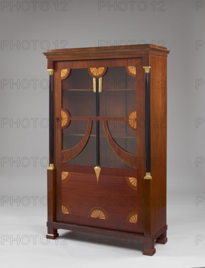 Library cabinet, maker, Léopold Méyet 1850–1912 author?;, from Méyet; Empire style; ornamented with arrows, columns, platter motif; pre-war, 1st quarter of the 19th century; possibly from Warsaw; furniture — storage cabinet; height 169 cm, width 107 cm, depth 40 cm., A wooden cabinet with glass-front doors standing on short square legs, showing a polished brown wood finish with lighter wood inlay semicircular fan motifs and curved inlay accents, vertical black columns on each side topped and base-trimmed with small gold-colored fittings, a gold-colored decorative pendant at the center of the glass panel, interior shelves visible through the glass, and a neutral gray background; visible colors include various shades of brown, black, gold, and gray.