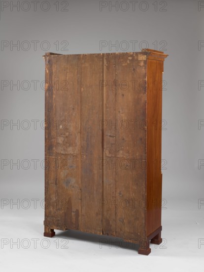 Library cabinet, empire style, arrows, columns, platter ornament, pre-war, 1st quarter of the 19th century, Warsaw, furniture, storage furniture; height 169 cm, width 107 cm, depth 40 cm., A tall wooden cabinet viewed from the back showing a large vertical wooden panel with visible grain, knots, scratches, and worn areas in brown tones, supported by a base with blocky feet in a darker brown; the cabinet's right side shows a smoother, polished wood surface in warm reddish-brown, and the top edge has a small overhanging molding in similar reddish-brown; the background is a smooth neutral gray gradient and the floor is a pale gray.