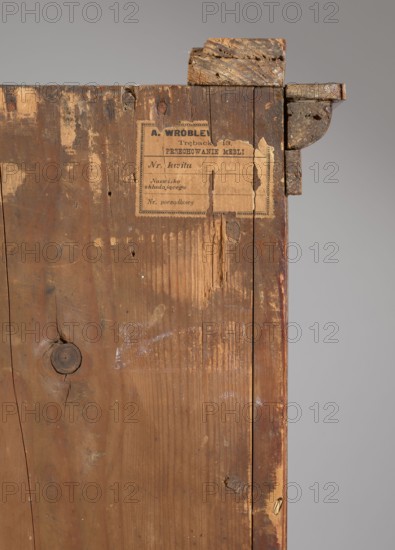 Library cabinet, empire style, 1st quarter of the 19th century, Warsaw, furniture, storage furniture; materials/ornament: arrows, columns, platter; dimensions: height 169 cm, width 107 cm, depth 40 cm., A vertical section of an old wooden panel with visible grain, knots, cracks, scratches, and worn edges, showing a torn paper label partially affixed near the top and a rough metal bracket attached at the upper corner; the wood displays shades of brown with areas of lighter beige where the finish is worn, and the background is a neutral gray.