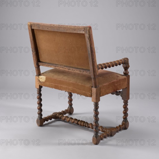 Armchair, Baroque, female busts, pre-war collection, ARMCHAIR, upholstered seat and backrest, with spirally turned frame elements and carved armrest supports. The legs end with glides-pads, with an H lacing at the bottom; the front legs are additionally fastened with a high link. At the joints of the footrest elements, ankles are formed, and the upper part of the front legs is elongated. The armrest supports are low, in the form of female busts mounted on turned balusters. Wide, rectangular seat and backrest; seat with secondary upholstery on springs; slightly reclined backrest, with a clearance, with turned side rails in the lower part. The whole upholstered with cherry plush covering the frame and backrest rails, finished with upholstery tape at the bottom of the backrest. 1640–1660, France. Seating piece furniture. Height 84.5 cm; width 61 cm; depth 51 cm., A wooden chair viewed from the back and slightly to one side with a worn brown upholstered seat and back; the upholstery shows ...