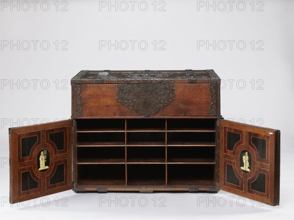 Cabinet, pre-war, grotesques ornament, fantastic animals, Hermes mythology, male figures, wickerwork ornament, fantastic figures, plant tendril. Cuboid cabinet with a drawer at the top and double doors below, richly decorated with openwork engraved fittings with grotesque motifs. The door leaves are veneered on the inside with rosewood and ebony in geometric arrangements, divided by inlaid bone veins, with figures in engraved bone, signed — Hippocrates on the left, Hermes on the right. Behind the door are twelve compartments. Drawer and door locks fitted with engraved steel plates; drawer lock blocks the left door leaf. Steel hinges. Late 17th century, Northern Germany, Poland . Piece of furniture, storage piece; height 70.1 cm, width 89.2 cm, depth 55.9 cm., A wooden chest with a dark brown finish sits against a light gray background, its front decorated with ornate dark metal fittings and a central metalplate with elaborate scrollwork; the chest's top shows darker metal straps and co...