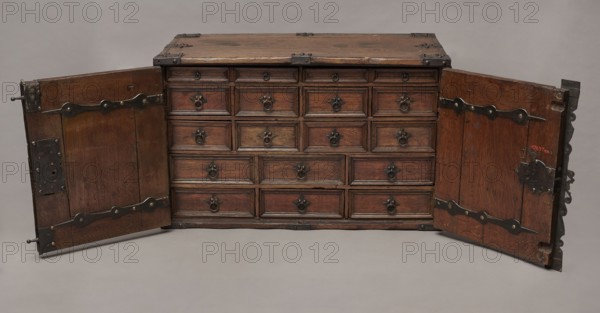 Cabinet, Baroque, 2nd quarter of the 17th century, Gdansk. A cuboid cabinet with a boarded body fitted with steel decorative strips, closed with double doors. Framed doors with single panels in the form of convex coffers glued at the mitre from richly profiled strips. Steel closing strip with a wavy outline, rectangular and trefoil-shaped angular fittings on the edges of the body, punched, fastened with headers. Inside: sixteen drawers arranged in five rows two in the top row, four in the two middle rows, three in the bottom rows. Drawer faces framed with profiled strips; three drawers in the middle row have numerous internal divisions. Walls connected with exposed diagonal dovetails; back joined to top and sides with dowels. Drawers with exposed diagonal dovetail joints; bottoms nailed. Side handles oval; drawer handles round with rosette-shaped plates. Hinges steel, strap-type with wavy outline. Both door leaves have steel mortise-and-apply locks with an additional hasp and covered k...