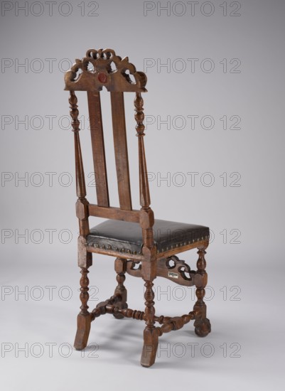Chair with turned and carved frame elements, padded seat and high backrest, supported by four turned legs fastened with turned links forming an H stretcher at the bottom, with an additional carved front board shell flanked by U-shaped acanthus leaves, topped with a closed crown. Front legs with carved feet in the form of a claw clamping a ball; rear legs angled outward with ankle feet. Backrest filled with two profiled slats, side frames in the shape of slender balusters, carved top board with decoration similar to the board connecting the front legs. Trapezoidal seat upholstered with leather covering the frame. Ornamentation: shells, plant decoration; motif: crown. Associated with the Camaldolese order/monastery. Baroque style, ca. 1700, possibly Gdansk. Seating furniture; height 124.7 cm, width 51.5 cm, depth 52 cm., A wooden chair with a tall back featuring carved decorative cresting and turned vertical supports, a dark leather upholstered seat with decorative studs along its edge, ...