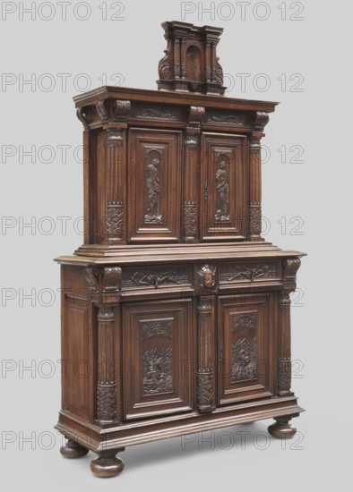 Cabinet by Jean Goujon ca. 1510–ca. 1565, co-creator, Renaissance style, ca. 1550, France. Two-part rectangular cabinet base and upper part with a front divided by three carved semi-columns; two-leaf doors; two drawers in the upper part of the base. Carved with architectural elements, fantasy animals, dolphins, tendrils, nymphs, and acanthus ornament. Storage furniture, height 191 cm, width 110 cm, depth 43 cm., A tall carved wooden cabinet with a dark brown finish occupies the image, standing on round bun feet and set against a light grey background. The piece has an ornate upper section with a small architectural pediment featuring carved scrolls and a recessed niche, and a larger lower section with a stepped projecting top surface. The cabinet doors and panels display detailed relief carvings including human figures, foliage motifs, and decorative mouldings, framed by fluted pilasters and carved capitals. A carved face motif appears on the frieze above the lower doors, and the wood ...