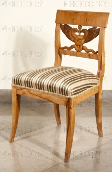 Chair, designed by Karl Friedrich Schinkel 1781–1841; maker, attributed to Paulina Sobolewowa ca. 1839–1919 in provenance. Biedermeier style, 1825–1835, possibly Poland. Biedermeier chair with a trapezoidal upholstered seat and a rectangular openwork backrest carved with a caduceus two winged snakes intertwined on a staff. Rectangular legs arched outward; back legs merge into the backrest frames. Straight frame. Seat upholstered in striped fabric. Backrest topped with a wide board; bottom is slatted. Seating furniture, height 84.3 cm, width 48.3 cm, depth 48.3 cm., A wooden chair with a light brown finish and carved decorative backrest sits on a pale gray tiled floor against a cream-colored wall; the seat is upholstered in a striped fabric featuring shades of beige, brown, and cream with a braided trim, the chair legs are curved and polished light brown wood, and a soft shadow is cast on the wall.