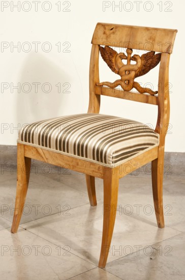 Chair, attributed to Karl Friedrich Schinkel 1781–1841 as designer author of the original, Biedermeier style, c. 1825–1835, Poland, seating furniture. Biedermeier chair with a trapezoidal upholstered seat and a rectangular openwork backrest carved with a caduceus two winged snakes intertwined on a staff. Rectangular legs arched outward; back legs merge into the backrest frames. Straight frame; seat upholstered in striped fabric. Backrest topped with a wide board; bottom is slatted. Height 84 cm, width 48 cm, depth 49 cm., A wooden chair with a carved backrest showing a symmetrical motif and wing-like shapes, a cushioned seat covered in a striped fabric, tapered wooden legs, and a light-colored wall and tiled floor in the background; visible colors include shades of brown, beige, cream, and a few darker brown tones.
