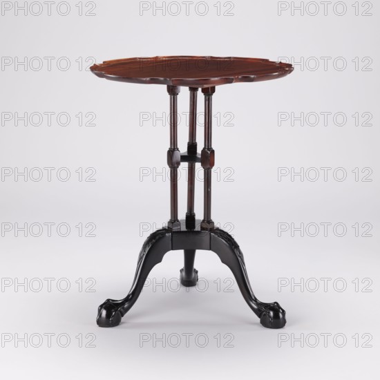Side table with round top and wavy profiled rim, supported by a shaft of three slender turned balusters fastened together and mounted on a carved tripod. Cabriole legs with feet in the form of a claw clasping a ball and strongly widened knees decorated with an acanthus leaf at the top, connected by a hexagonal cube with three concave sides. The balusters are fastened together midway by a shelf of analogous shape; the shaft balusters have angular cubes at the top and bottom and at the junction with the shelf. Animal paws, acanthus ornament., 2nd half of the 18th century, England. Furniture table. Height 55.1 cm; diameter 44 cm., A small round wooden table with a scalloped edge top in a reddish-brown color supported by three slender vertical supports that join at a central block, which sits on a tripod base with three curved legs carved with decorative elements and ending in paw-like feet; the legs and lower base are black, and the background is light gray.