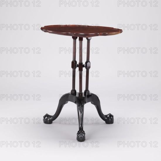 Table, SIDE TABLE with a round top with a wavy, profiled rim, supported by a shaft composed of three slender turned balusters fastened together and mounted on a carved tripod. Cabriole legs with feet in the form of a claw clasping a ball and strongly widened knees decorated with an acanthus leaf at the top, connected by a hexagonal cube with three concave sides. The balusters are fastened together midway by a shelf of analogous shape. The shaft balusters have angular cubes at the top and bottom and at the junction with the shelf. Features: animal paws, acanthus ornament. 2nd half of the 18th century, England. Furniture; table. Height 55.1 cm; diameter 44 cm., A wooden tripod table with a scalloped oval top in a reddish-brown finish supported by a set of three slender turned columns that are also reddish-brown, joined near their midpoints by small dark brown blocks, and resting on three curved black legs that end in carved paw-like feet; the background is light gray and white.