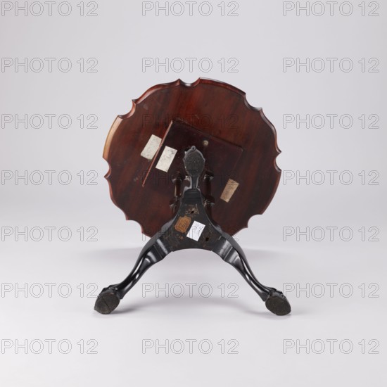 SIDE TABLE with a round top with a wavy, profiled rim, supported by a shaft composed of three slender turned balusters fastened together and mounted on a carved tripod. Cabriole legs with feet in the form of a claw clasping a ball and strongly widened knees decorated with an acanthus leaf at the top, connected by a hexagonal cube with three concave sides. The balusters are fastened together midway by a shelf of analogous shape. Shaft balusters with angular cubes at the top and bottom and at the junction with the shelf. Animal paws; acanthus ornament. 2nd half of the 18th century, England. Furniture, table. Height 55.1 cm; diameter 44 cm., A round wooden table top with a scalloped edge is shown flipped upright, revealing a reddish-brown polished surface with darker wood grain. The underside of the table top displays a small square wooden bracket and a few rectangular lighter patches. A carved tripod base in glossy black supports the top, with three curved legs ending in rounded feet tha...