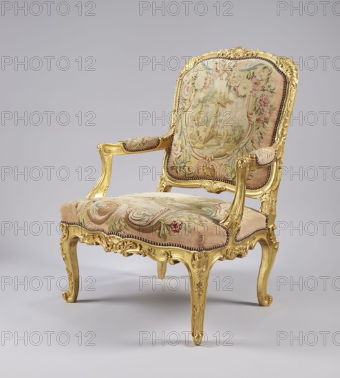 Armchair by Jean-Baptiste Oudry 1686–1755 and unknown co-creator; carved, gilded, and upholstered Rococo armchair in the à la reine style, 3rd quarter of the 18th century, France. Tapestry upholstery possibly Aubusson or Beauvais featuring scenes from Jean de La Fontaine's fables framed with flowers: backrest depicts an insect nest against a landscape illustration of The Hornets and the Bees; seat depicts a wolf and a lamb by a stream The Wolf and the Lamb. Motifs include wolves, lambs, insects, flowers, bees, shells, acanthus leaves, rocaille elements, and bunches of roses. Cabriole/S-shaped legs with acanthus-shaped volute feet; volute-topped recessed armrests on U-shaped supports; roughly rectangular slightly concave backrest with convex top and rounded corners; trapezoidal seat with slightly bulging frames. Function: seating piece. Dimensions: height 106.5 cm, width 77.5 cm, depth 79.4 cm., A decorative armchair with a carved gilded wooden frame and curved legs upholstered in patte...