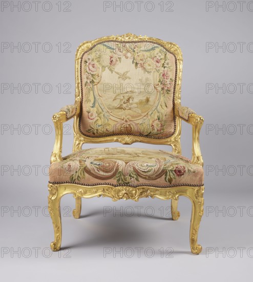 Armchair, possibly upholstered with Aubusson or Beauvais fabric, attributed to Jean-Baptiste Oudry 1686–1755 for illustrations for fairy tales; decorations include flowers, vultures, wolves, birds, pigeons, wells, animals, shells ornament, Rococo ornament and style; references Jean de la Fontaine 1621–1695 Fairy Tales; cabriole legs; 3rd quarter of the 18th century, France; seating piece of furniture; height 107.3 cm, width 76.8 cm, depth 77.5 cm., An ornate upholstered armchair with a carved gilded wooden frame and cabriole legs, featuring floral and scroll tapestry fabric in shades of beige, pink, green, soft blue, and muted gold; the backrest and seat show decorative floral and scroll motifs with a central pastoral scene in pale beige tones, and the armrests are padded with matching fabric; the frame has carved acanthus and shell details in bright gold, with a row of dark round decorative nails along the edge of the upholstery, all set against a neutral light gray background.