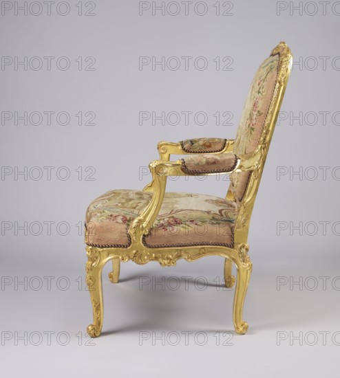 Armchair, created in the 3rd quarter of the 18th century, France. Upholstery fabric possibly Aubusson or Beauvais. Seating piece of furniture with cabriole legs; height 107.3 cm, width 76.8 cm, depth 77.5 cm. Decoration includes illustrations for fairy tales after Jean de la Fontaine, flowers, vultures, wolves, birds pigeons, wells, animals, shells and Rococo ornament and style. Designer: Jean-Baptiste Oudry 1686–1755 attributed for original illustrations; maker, origin/provenance unknown., A gilded wooden armchair with carved scrollwork and cabriole legs upholstered in a floral-patterned fabric; the frame is gold in color and the upholstery shows a palette of pink, green, beige, and cream with floral motifs; a few decorative nailhead trims in a dark tone outline the seat and arm pads; the chair is shown from the side against a neutral pale gray background.