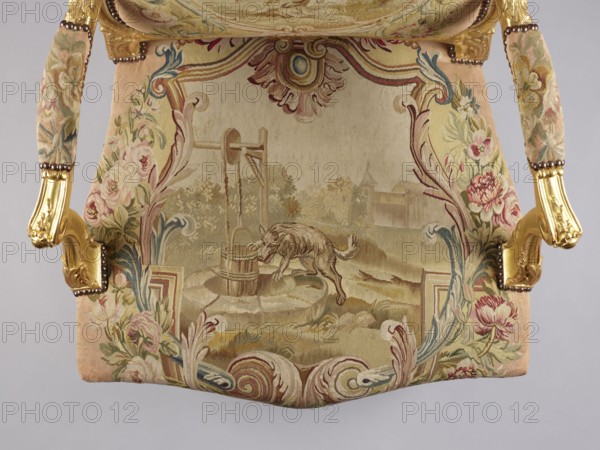 Armchair, maker, upholstery attributed to/to designs by Jean-Baptiste Oudry 1686–1755 and/or after illustrations for Jean de La Fontaine's fairy tales Jean de La Fontaine, 1621–1695. Decor: flowers, vultures, wolves, birds including pigeons, wells, other animals; Rococo ornamentation and shells; cabriole legs. 3rd quarter of the 18th century, France Aubusson and/or Beauvais upholstery. Seating piece of furniture; height 107.3 cm, width 76.8 cm, depth 77.5 cm., An upholstered chair seat and part of its arms are visible, showing a woven fabric with an ornate scene and floral borders; the fabric displays muted tones of beige, cream, pink, green, blue, and gold, with a central pastoral image that includes an animal near a well and surrounding foliage, and the armrests feature gilded carved wood with brass or bronze nailhead trim and floral-patterned fabric covers in matching colors.