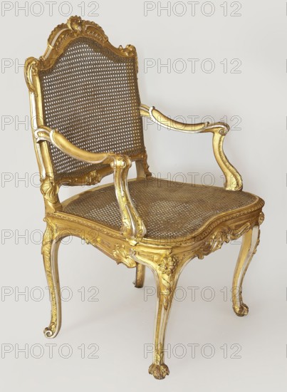 Armchair, origin possibly Italy or Southern Germany, Rococo style, 3rd quarter of the 18th century. Carved, gilded and silver-plated frame originally white lacquered with gilded and silver-plated elements; seat and backrest woven with rattan thread. S-shaped legs with volute feet front ones shaped like a leaf. Wavy-cut lower frame; trapezoidal applied-seat frame with rounded front corners. Backrest shaped like a lying rectangle topped with an arch, straight with the back and slightly tilted back, with a clearance. S-shaped armrests and supports connected by a volute. Profiled frame elements with edges highlighted by a half-cylinder, decorated with acanthus leaves, rocaille motifs middle of upper and lower backrest frames and front frame, and floral sprigs on the knees of the legs. Seating furniture; height 101.6 cm, width 73 cm, depth 66.5 cm., A decorative chair with a carved frame finished in gold and a woven cane seat and back, featuring curved armrests and ornate floral and scroll ...