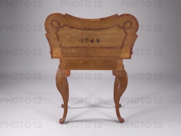 Game table, baroque style, possibly made 1748. Rectangular frame with an extendable tabletop that when unfolded is nearly a rectangle with rounded corners and concave sides; supported by four cabriole, smooth legs. Legs are strongly curved and widened at the knees with a ridge defining symmetry and widened feet so-called vipers. The frame extends to support the foldable top plate; wide, flat frame. The tabletop is inlaid on both sides with a ribbon ornament that frames the edges and divides the central field, with loops in the rounded corners, and the date 1.7.4.8 in black on the outside. On the upper surface of the unfolded tabletop, in the center of each side, are recesses for tokens and stones. Construction includes steel scissor hinges. Northern Germany attributed. Piece of furniture — table. Height 73 cm; width 90 cm; depth 87.3 cm., A wooden piece of furniture viewed from the back with a curved, decorative top and carved, cabriole-style legs, showing wood grain and darker inlaid ...