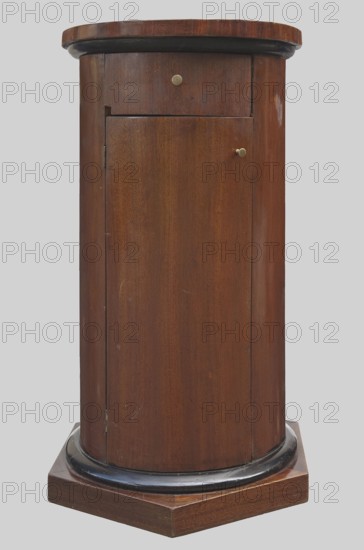 Cabinet-pedestal, message provenance, columns architectural motif, classicism and empire styles, early 19th century, possibly Northern Germany or Poland, piece of furniture storage, height 84 cm, diameter 46.5 cm., A cylindrical wooden cabinet in brown tones with a darker brown or black rounded trim near the top and base, sitting on a brown hexagonal wooden platform; the cabinet has a small round metal knob on a small top panel and another small round metal knob on a larger front door panel, with visible wood grain and a few small scuffs and lighter marks on the surface, set against a plain light gray background.