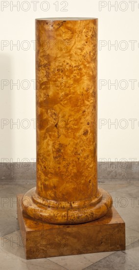 Pedestal, provenance unknown, Biedermeier style. PEDESTAL in the shape of a column with a smooth shaft, with a cylindrical base on a square plinth. The whole is veneered with birch burl. The shaft is made of eight boards glued together at an angle from the front. The four plinth boards are connected with diagonal dovetails. The shaft is fastened to the plinth with four screws and additionally mounted on a centrally placed peg. 1825–1835, Poland, furniture. Height 112 cm, width 44.7 cm, depth 45 cm., A tall cylindrical wooden column with a glossy burl wood grain pattern in shades of golden brown and amber stands on a round stepped wooden base of the same material, which sits on a square wooden plinth; the floor around it is stone in muted gray tones and the wall behind is a pale off-white.