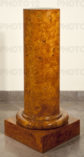Pedestal, creator, provenance unknown. Biedermeier style. Pedestal in the shape of a column with a smooth shaft, a cylindrical base, on a square plinth. Entire piece veneered with birch burl. Shaft made of eight boards glued together at an angle from the front. Four plinth boards connected with diagonal dovetails. Shaft fastened to the plinth with four screws and additionally mounted on a central peg. Dated 1825–1835. Likely Poland. Furniture. Height 111.5 cm; width 44.5 cm; depth 44.5 cm., A tall cylindrical wooden column with a glossy, richly grained surface in warm amber and honey brown tones standing on a rounded wooden base and a square plinth of matching wood grain, set on a gray stone floor against a pale cream wall.