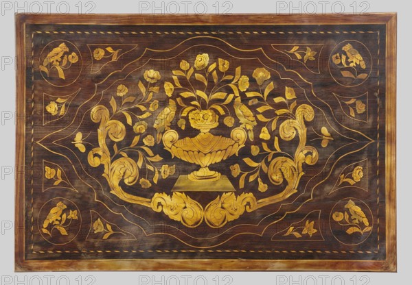 Table, birds, plant ornament, vases with flowers, classicism, late 18th century, Netherlands, furniture table; height 73 cm, width 89 cm, depth 56 cm, A rectangular wooden panel with a dark brown background and a lighter brown outer frame, decorated with an ornate golden floral inlay; at the center is a golden urn or vase holding a bouquet of stylized flowers and leaves, flanked by a pair of golden birds perched on the vase; surrounding the central motif are flowing golden scrolls, curving acanthus-like leaves, and a scattering of small floral elements; the corners feature circular medallions each containing a bird and floral motif, and the border area shows a repeating twisted rope or braided pattern in gold; visible colors include dark brown, various warm golden tones, and lighter brown in the frame.
