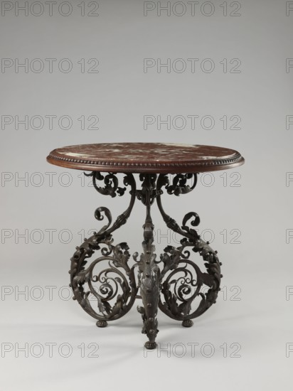 Table, Szewczykowski, Roman 1849–1901 author, plant decorations, plant twig, astragal, in the form of three S-shaped pieces, 1890s, Warsaw, furniture, table, height 80.7 cm, diameter 84 cm, A round table with a brown and white mottled top and a carved brown wooden rim stands on an ornate dark metal base; the base features a group of scrolling, leafy, and curling decorative elements with a dark bronze or black finish and small rounded feet, and the overall color palette includes brown, white, dark bronze or black, and hints of lighter gray from reflections.