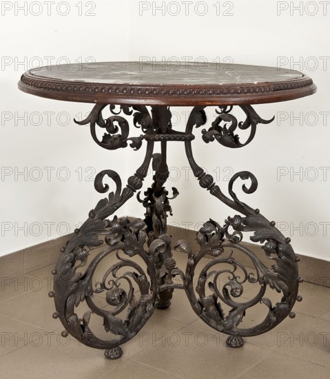 Table, Szewczykowski, Roman 1849-1901 creator, plant decorations, plant twig, astragal, in the form of three S-shaped pieces, 1890s, Warsaw, furniture, table, height 80.7 cm, diameter 84 cm, A round table with a dark brown wooden rim and a grayish top sits against a light beige wall and on a tan tiled floor, supported by an ornate dark metal base with intricate scrolling leaves and floral motifs forming a group of curving legs, with visible small round decorative knobs and carved details in dark brown and black tones.