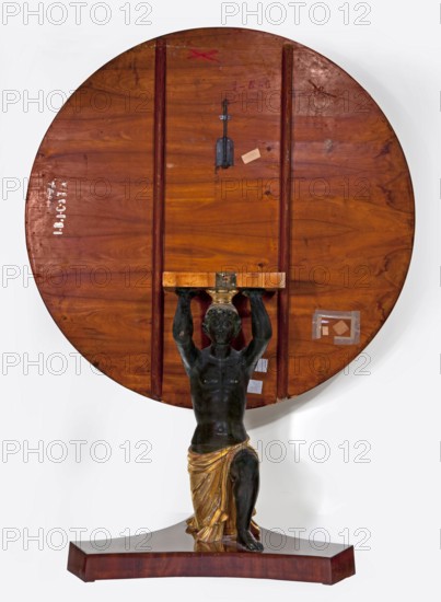 Table, empire style, furnishings of representative buildings of the Republic of Poland 1918–1939, Administration of the Imperial Palaces in Warsaw 1815–1915, male figures, dated 1816–1825, place of origin possibly Warsaw or Vienna, furniture; table, height 81 cm, diameter 110 cm, A large round wooden tabletop with a rich brown and reddish grain rests upright, showing the underside with a few vertical wooden supports and a small metal latch; attached near the bottom center is a dark greenish-black sculpted figure of a kneeling man holding the tabletop above his head, the figure wearing a draped garment painted in gold tones; the whole piece stands on a curved wooden base that shares the brown and reddish wood hues, and there are a few small pale patches and adhesive squares visible on the underside.
