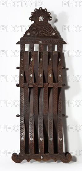 Chair, rosettes and geometric ornament, Kufi script. Scissor chair with footrest, seat, and backrest composed of five slatted elements; bas-relief and inlaid with bone with geometric and plant patterns. Supports connected to each other and to the seat with wooden pins enabling folding and unfolding, terminating at the bottom with decoratively carved runners. Backrest topped with a wide board with a wavy outline resembling a triangle. Slats decorated on front and sides with stylized plant patterns; backrest topped with symmetrical geometric decoration and a bas-relief Arabic verse. Pins topped with turned knobs. Mid-19th century 19th century, Syria place of origin, piece of furniture, seating piece; height 107 cm, width 46 cm, depth 47.5 cm., A tall carved wooden object with a dark brown finish featuring ornate repeating carved patterns and floral inlay accents in a lighter color, shown against a plain light background. The top has a scalloped decorative crest with circular cutouts and ...