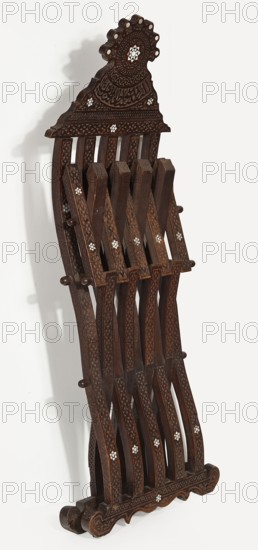Chair, rosettes ornament, geometric ornament, Kufi script. Scissor chair with footrest, seat, and backrest composed of five slatted elements; bas-relief and inlaid with bone with geometric and plant patterns. Supports are connected to each other and to the seat with wooden pins enabling the structure to be folded and unfolded, terminating at the bottom with decoratively carved runners. Backrest is topped with a wide board with a wavy outline resembling a triangle. Slats are decorated on the front and sides with stylized plant patterns; the backrest is topped with symmetrical geometric decoration and a bas-relief Arabic verse. Pins are topped with turned knobs. Mid-19th century 19th century, Syria place of origin, piece of furniture, seating piece. Dimensions: height 107 cm, width 46 cm, depth 47.5 cm., A carved wooden folding stand with a dark brown finish featuring a decorative crest at the top and curving slats that interlock to form a lattice; the wood shows carved geometric and flo...