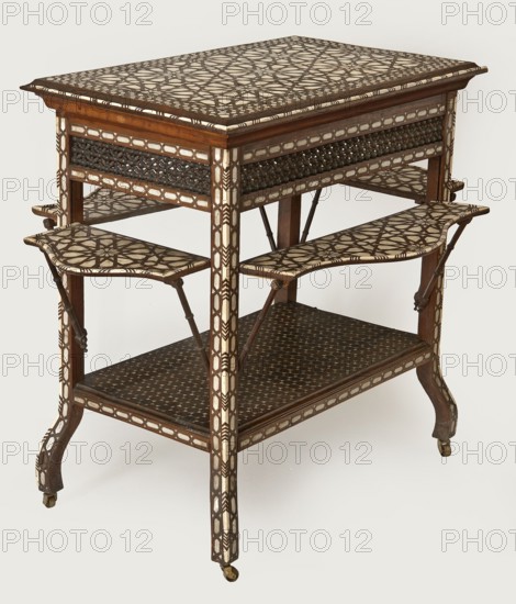 Side table. Rectangular side table with drop-down desks on each side and a bottom shelf, inlaid with mother-of-pearl and bone in geometric and plant patterns. Straight, angular legs S-shaped at the bottom, topped with brass rollers. Wide, openwork frame filled with a dense lattice of turned elements mashrabiya. Tabletop, desks, frame, and shelf edge decorated with geometric braid forming rosettes on the surfaces. Shelf covered with stars with hexagonal fields in the center, forming a network. Desk supports imitate bamboo. 2nd half of the 19th century. Syria, furniture, table. Dimensions: height 82 cm, width 100 cm, depth 75 cm., A wooden table with decorative inlay showing a geometric lattice pattern in cream and dark brown on the top, sides, and two folding shelves, featuring a darker wood frame with carved and beaded detailing, slender legs with cream inlay accents, small caster wheels at the feet, and a lower shelf with a repeating circular motif in dark brown and cream.