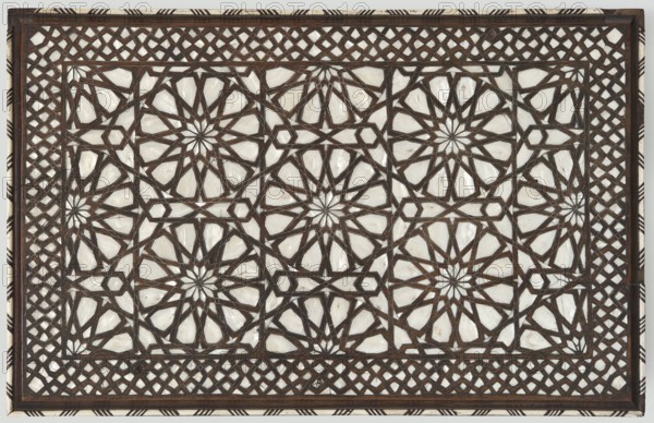 Side table, noted; geometric ornament, rosettes, stars, mashrabiya. Rectangular side table with drop-down desks on each side and a shelf at the bottom, inlaid with mother-of-pearl and bone in geometric and plant patterns. Straight, angular legs, S-shaped at the bottom, topped with brass rollers. Wide, openwork frame filled with a dense lattice of turned elements mashrabiya. The tabletop, desks, frame edges, and shelf edge are decorated with geometric braid forming rosettes on the surfaces. The shelf is covered with stars with hexagonal fields in the center, forming a network. The desk supports imitate bamboo. 2nd half of the 19th century, probably Syria. Furniture; table. Dimensions: height 82 cm, width 100 cm, depth 75 cm., A rectangular decorative panel with an intricate geometric lattice of interlocking star and polygon shapes, featuring a dark brown linear framework over light, creamy white inlay; the design repeats across the surface with a border of diagonal woven lattice and a n...