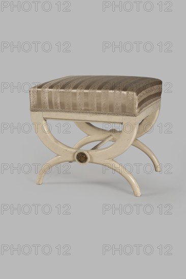Stool, origin unknown, Empire style, rosette ornament. Rectangular stool with a black painted frame, upholstered, supported on the longer sides by crossed supports composed of two arched squares fastened in the middle and connected with a turned, simple link. At the intersections of the crosses are turned plaques with round fittings of embossed gilt brass sheet, each with a central rosette and a tendril on the edge. Padded seat with high sides, upholstered with non-original green striped fabric alternating grosgrain and satin. c. 1810, Central Europe. Furniture; seating piece. Dimensions: height 40 cm, width 43.2 cm, depth 35.3 cm., A small upholstered stool with a striped fabric seat in shades of beige and light brown, sitting on a curved wooden frame painted cream; the seat has a subtle patterned texture and a decorative trim along its lower edge, and the frame features intersecting curved legs forming an X-shape with a round decorative medallion at the center, all shown against a pl...