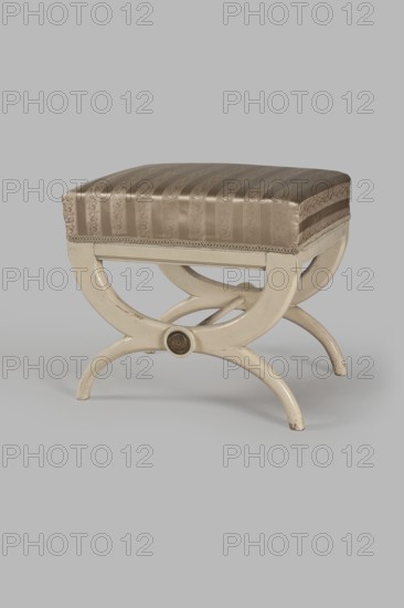 Stool, origin unknown, Empire style. Rectangular stool with a black painted frame, upholstered, supported on the longer sides by crosses composed of two arched squares fastened in the middle and connected with a turned simple link. At the intersections of the crosses are turned plaques with round fittings of embossed gilded brass sheet, featuring a central rosette and a tendril on the edge. Padded seat with high sides, upholstered with non-original green striped fabric alternating grosgrain and satin. c. 1810, Central Europe. Furniture — seating piece. Height 40 cm; width 43.2 cm; depth 35.3 cm., A small upholstered stool with a rectangular padded seat covered in a shiny striped fabric in shades of brown and beige, the fabric showing a subtle floral pattern and decorative trim along the lower edge; the base is a painted wooden X-shaped frame in an off-white or cream color with curved legs and a circular carved medallion at each end, showing slight wear and scuff marks on the paint.