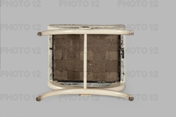 Stool, origin unknown, Empire style, rosette ornament. Rectangular stool with a black painted frame, upholstered, supported on the longer sides by crosses composed of two arched squares fastened in the middle and connected with a turned, simple link. At the intersection of the crosses are turned plaques with round fittings of embossed, gilded brass sheet, with a central rosette and a tendril on the edge. Padded seat with high sides, upholstered with non-original green striped fabric alternating grosgrain and satin. Circa 1810, Central Europe. Furniture, seating piece; height 40 cm, width 43.2 cm, depth 35.3 cm., An overturned stool with curved tubular legs and a central support, showing its underside with woven fabric webbing; the stool frame and legs are off-white or cream with areas of wear exposing darker spots, and the webbing is brown; the background is a flat light gray.