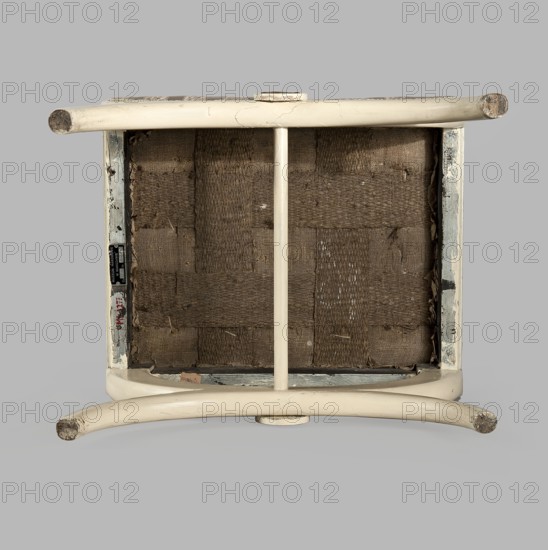 Stool, origin unknown, Empire style, rosette ornament. Rectangular stool with a black painted frame, upholstered, supported on the longer sides by crosses composed of two arched squares fastened in the middle and connected with a turned, simple link. At the intersection of the crosses are turned plaques with round fittings of embossed gilded brass sheet, with a central rosette and a tendril on the edge. Padded seat with high sides, upholstered with non-original green striped fabric alternating grosgrain and satin. c. 1810, Central Europe. Furniture, seating piece. Dimensions: height 40 cm, width 43.2 cm, depth 35.3 cm., An upside-down wooden stool or small chair with a cream or off-white painted frame showing scuffs and worn areas, a woven seat underside made of coarse brown fibers in a grid pattern, a central vertical support painted the same cream color, rounded legs with exposed worn ends, and a neutral light gray background.