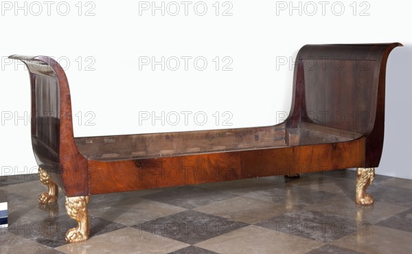 Bed, empire style, mahogany, with S-shaped outward-curving pediments of slightly different heights and straight side boards, supported by four carved, black-painted and partly gilded legs in the form of animal paws. Frame pediments with single panels, connected to the sides with hooks. Inscription/notation: unknown, Date: 1815–1825. Place of origin: Warsaw. Function: piece of furniture. Dimensions: height 104.5 cm, width 231.5 cm, depth 98 cm., A wooden sleigh-style bed frame with high curved headboard and footboard, showing a polished brown wood grain finish, sits on a checkered stone floor. The bed frame has carved ornamental legs shaped like pawed animal feet with a gilded gold color, and the interior wood of the frame appears lighter and unfinished compared with the darker outer panels. The floor tiles show a mix of beige, gray, and dark gray tones, and the background is a plain pale wall. The visible colors include brown, dark brown, gold, beige, gray, and pale off-white.