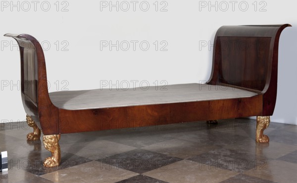Bed, empire style, animal paws, mahogany, with S-shaped outward-curving pediments of slightly different heights and straight side boards, supported by four carved, black-painted and partly gilded legs in the form of animal paws. Frame pediments with single panels, connected to the sides with hooks. 1815–1825, Warsaw, piece of furniture. Height 104.5 cm; width 231.5 cm; depth 98 cm., A wooden sleigh-style bed with curved headboard and footboard in a dark brown finish, a light-colored mattress or cushion on the frame, ornate gold-painted carved feet shaped like animal paws, a polished stone or tile floor in shades of gray and brown, and a plain light gray wall behind the bed; visible colors include dark brown, light beige, gold, gray, and brown.