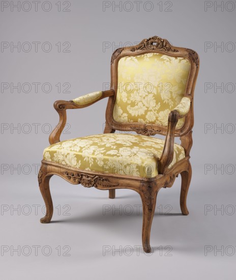 Armchair, rococo style, carved and upholstered seat, backrest and armrests, on four cabriole legs ending in turned-out slipper feet, arching into a wavy-cut slightly bulging frame. Seat cushion recessed into the frame rebate, on straps. S-shaped armrests and supports. Backrest disc-shaped, nearly rectangular, tilted outward, with a clearance. Profiled frame elements enriched with carving rosettes on the knees of the legs; rocaille motif framed by a plant twig and a bird's wing on the front frame and horizontal backrest frames. Tenon joints reinforced with dowels; re-painted with gold paint. 3rd quarter of the 18th century, probably Italy. Piece of furniture — seating piece. Height 92 cm, width 70 cm, depth 64 cm., A wooden armchair with carved details and a warm brown finish features upholstered seat, back, and arm rests covered in patterned yellow and cream fabric; the chair has curved legs, decorative floral carvings on the frame, a cushioned seat and back with visible floral motifs,...