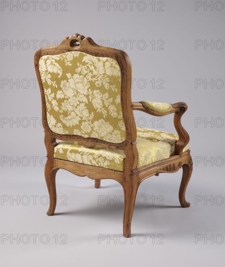 Armchair, rococo style, carved and upholstered seat, backrest and armrests, on four cabriole legs ending in turned-out slipper feet, arching into a wavy-cut slightly bulging frame. Seat cushion recessed into the frame rebate on straps. S-shaped armrests and supports. Disc-shaped, nearly rectangular backrest tilted outward with a clearance. Profiled frame elements enriched with carving rosettes on the knees of the legs; on the front frame and horizontal backrest frames a rocaille motif framed by a plant twig and a bird's wing. Tenon joints reinforced with dowels; re-painted with gold paint. 3rd quarter of the 18th century, Italy, seating piece, height 92 cm, width 70 cm, depth 64 cm., An upholstered wooden armchair shown from the back and slightly to the side with a carved wooden frame in a medium brown tone and padded seat, back, and armrest covered in a patterned yellow fabric with lighter floral motifs; the chair has curved legs and scrolled arm supports, visible wood grain and small...
